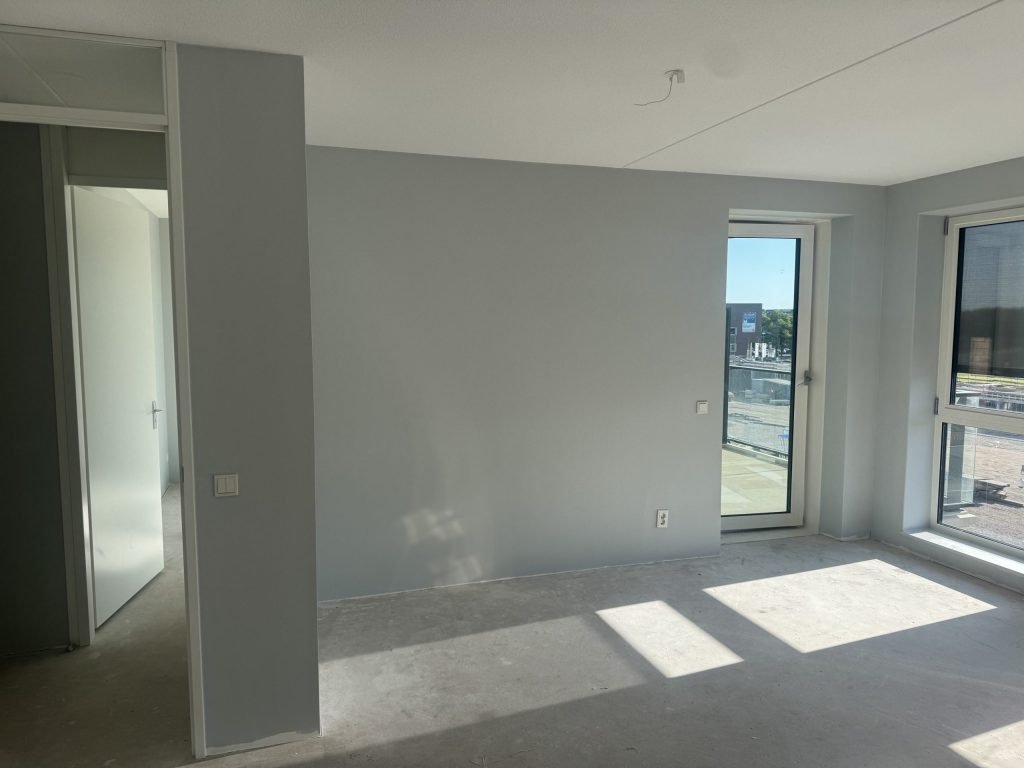 Kamer met lichtgrijs geverfde wanden en een deur naar het balkon, zon op de betonvloer