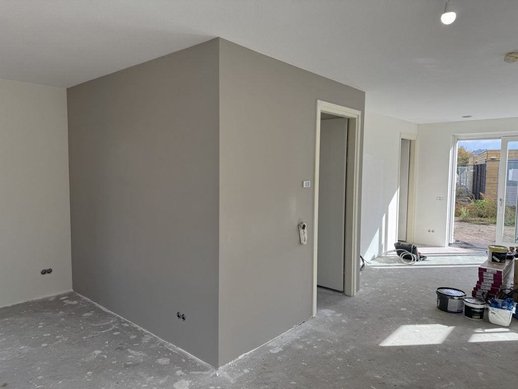 Nieuwbouwwoning met een pas geverfde grijze wand naast een deuropening en verfemmers op de vloer