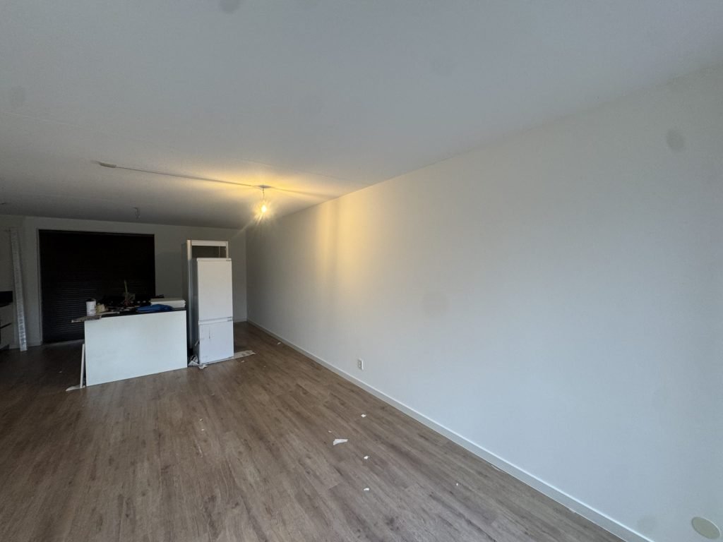 Lange, strak afgewerkte witte wand in een woonkamer met een keukenblok op de achtergrond